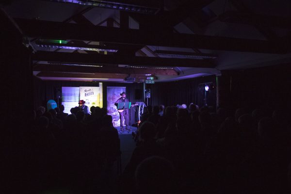 Photo of people seated in a darkened room listening to a man playing guitar and singing.