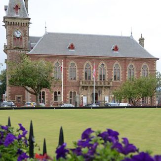 Photo of external view of two story Victorian building with clock tower. Bowling green and flowers in the foreground.